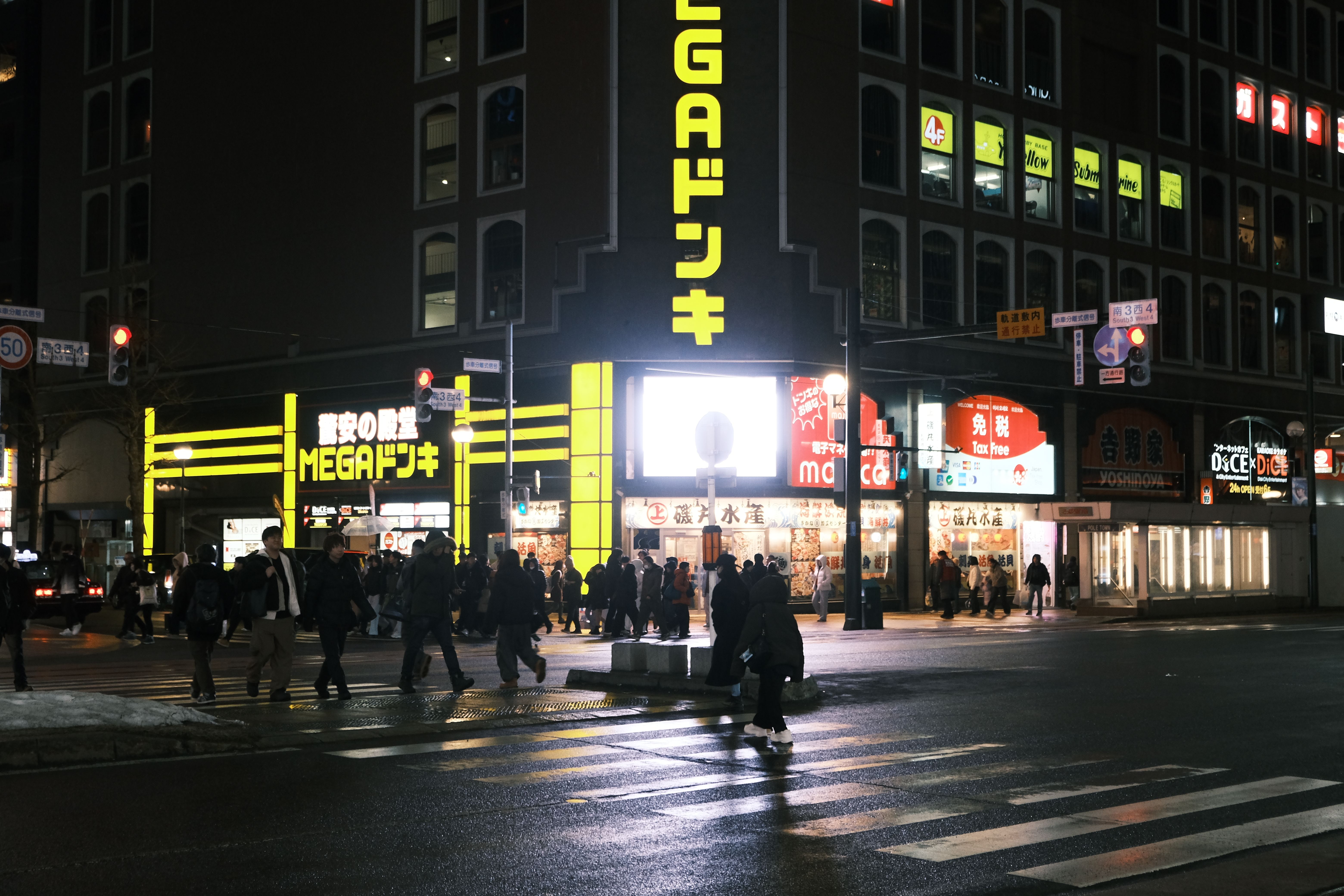 Busy nighttime street in Sapporo, Hokkaido with lit storefronts and crossing pedestrians.