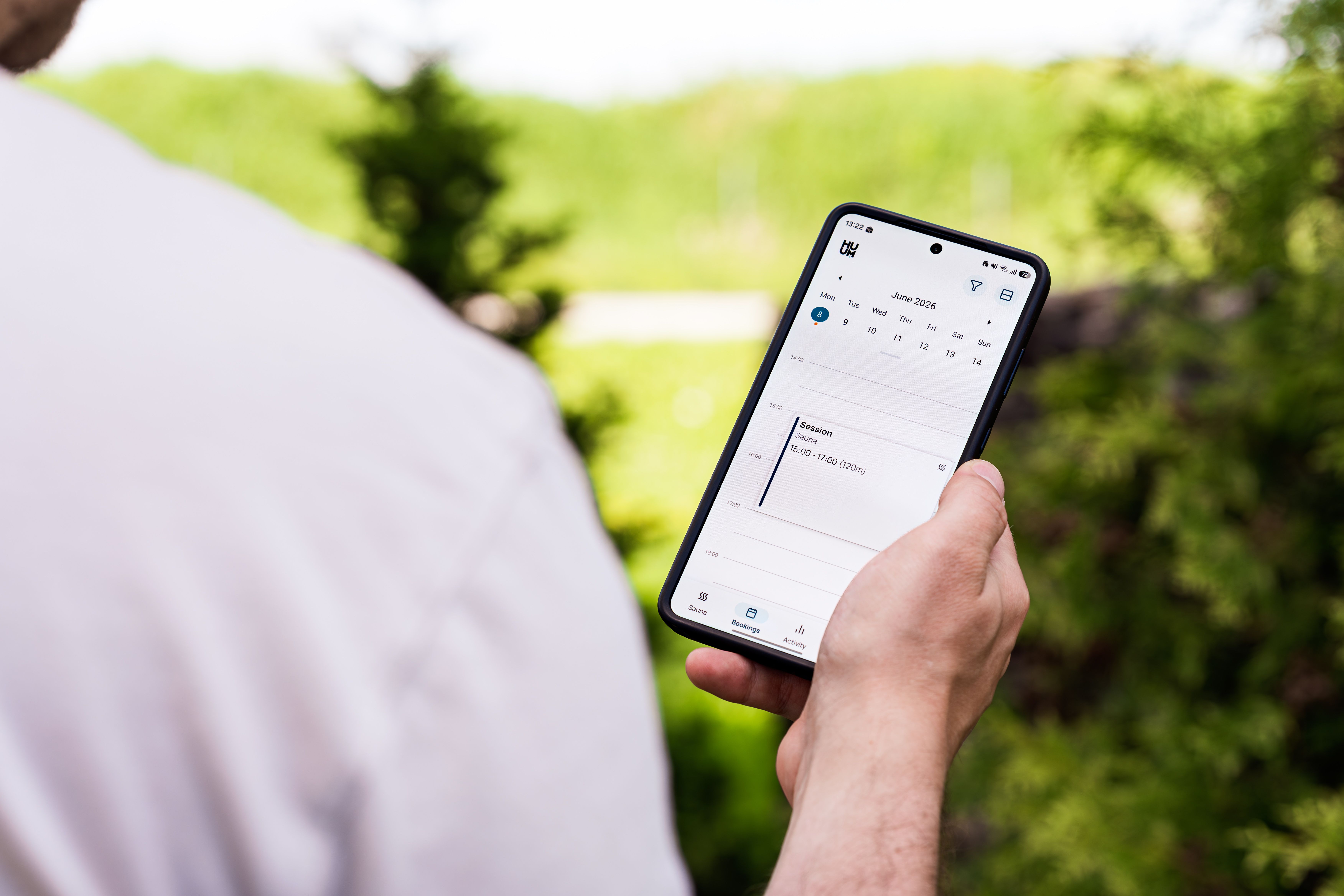 A person using a sauna booking app on a smartphone outdoors, surrounded by greenery.