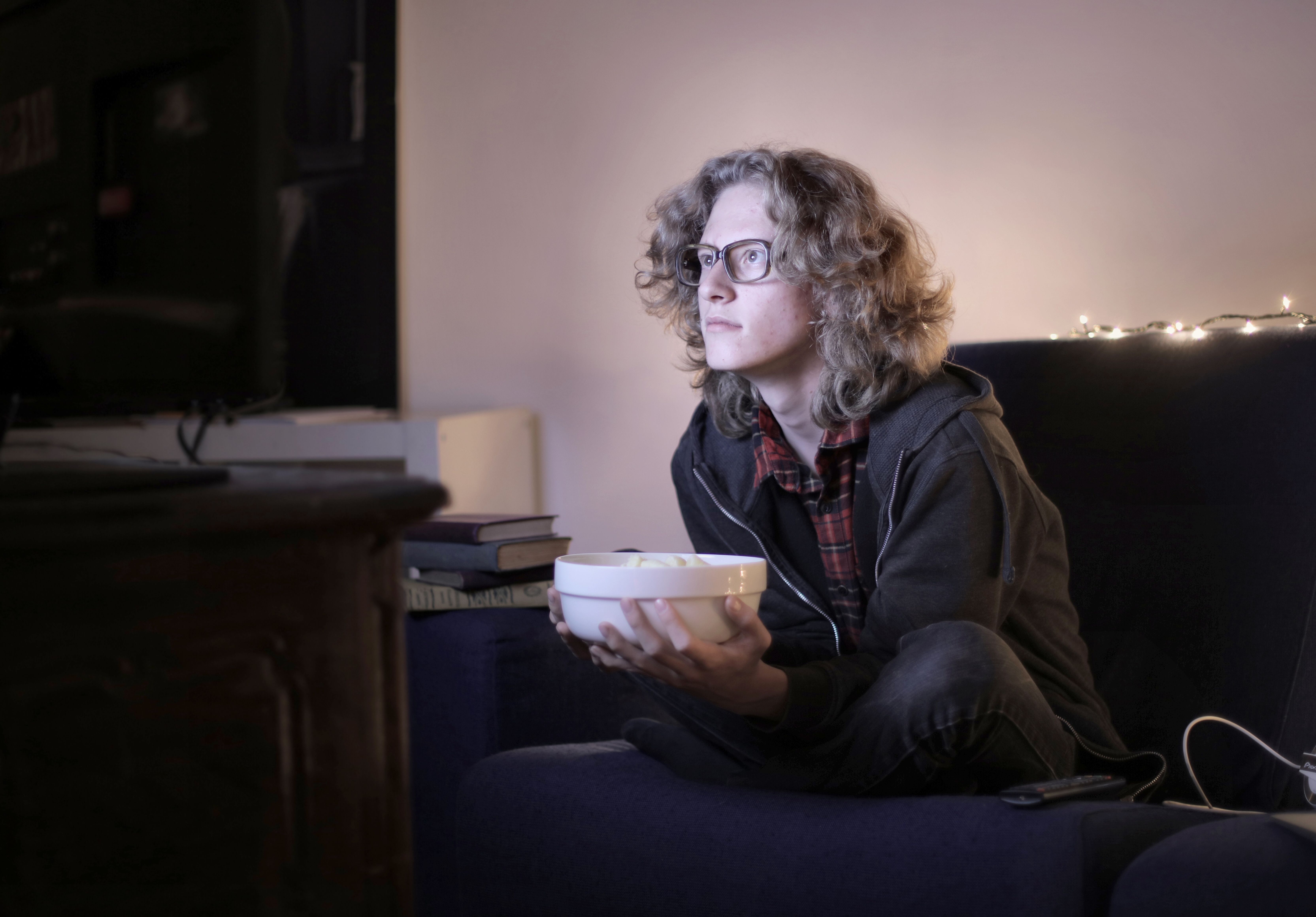 Concentrated male teenage with long hair in casual wear and eyeglasses sitting on couch with bowl of popcorn and watching interesting movie while resting in cozy living room at home