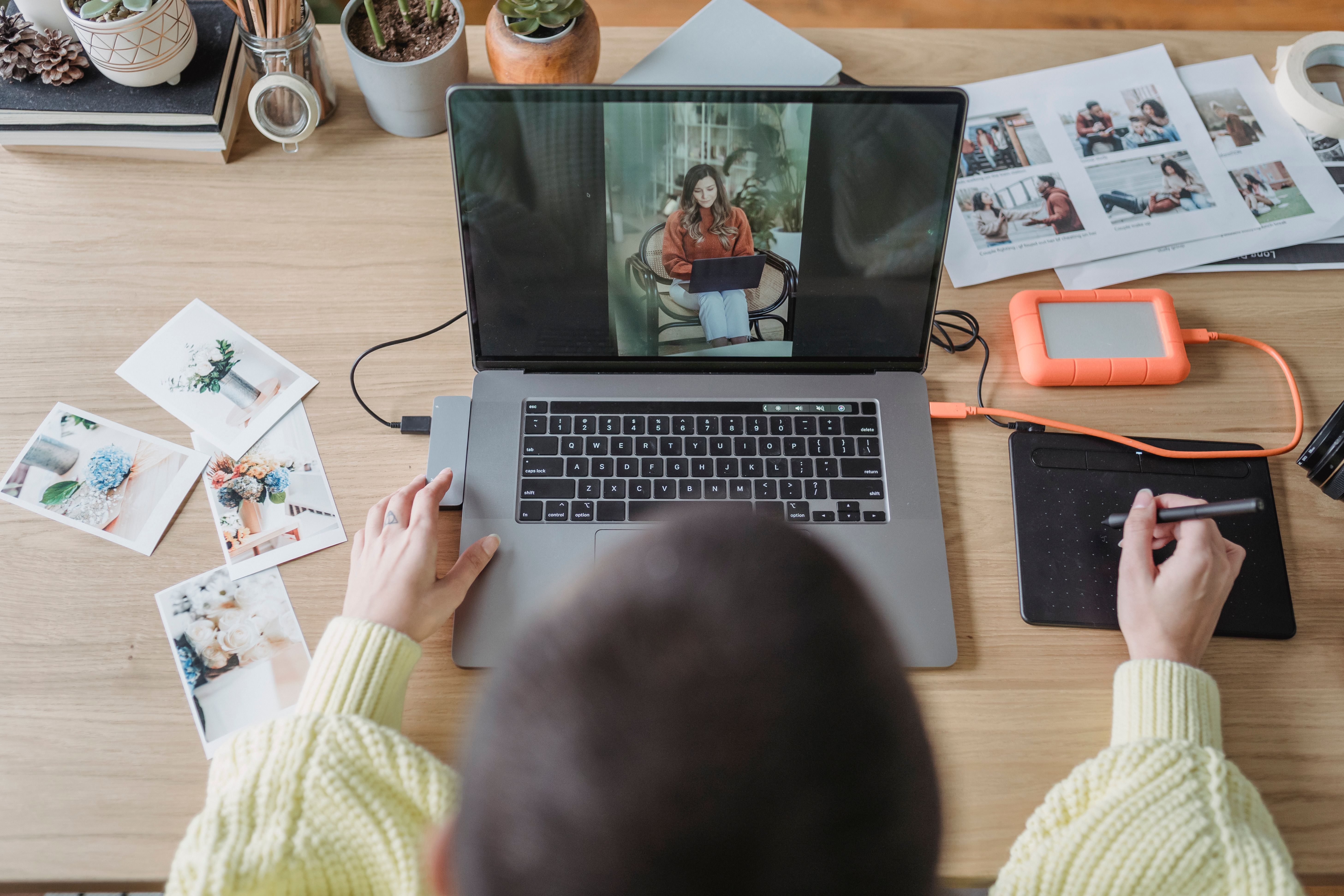 From above of crop faceless female photographer drawing on graphic tablet while editing photos on laptop at table with printed photos