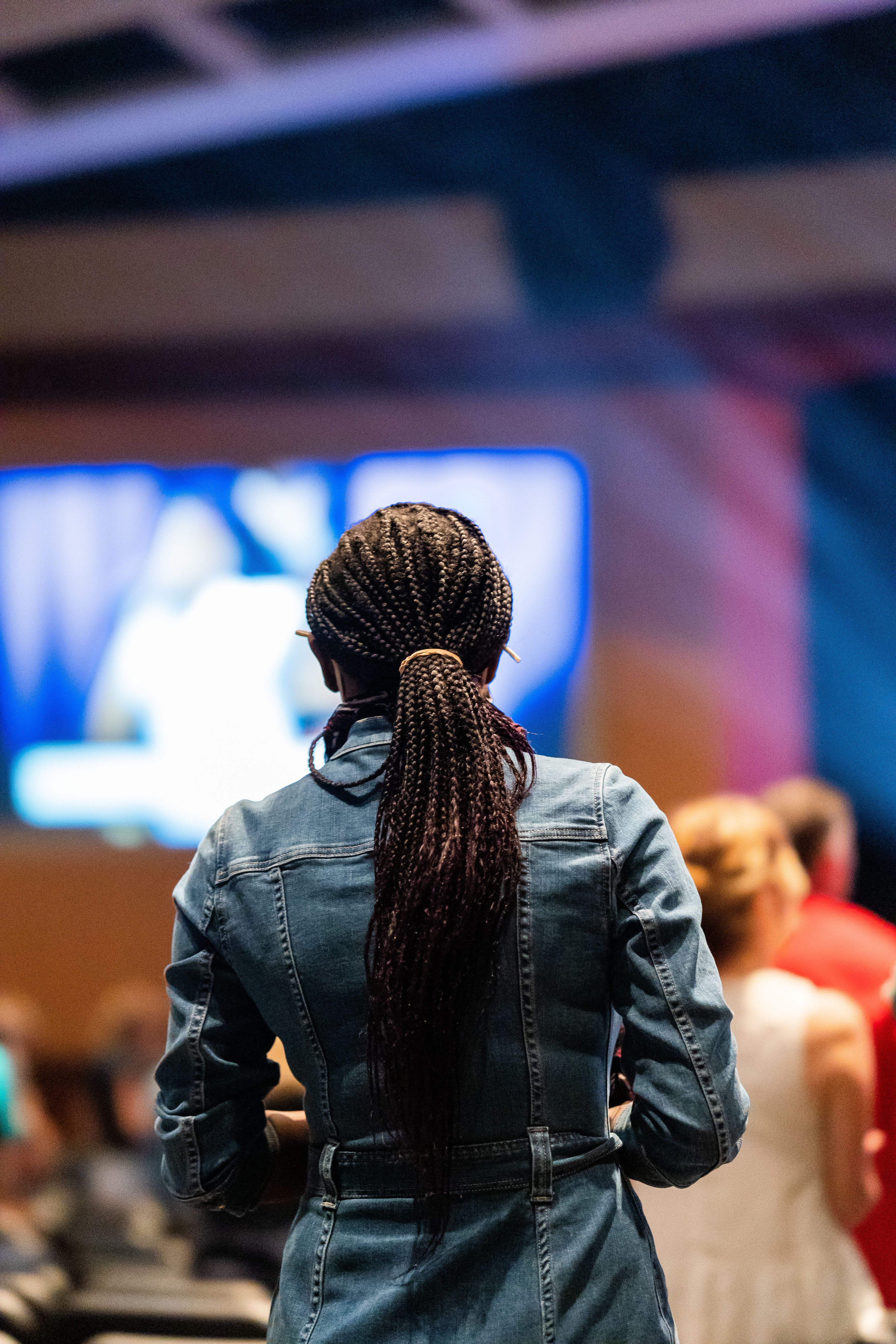Back view of unrecognizable female viewer with Afro braids enjoying live performance against screen and people