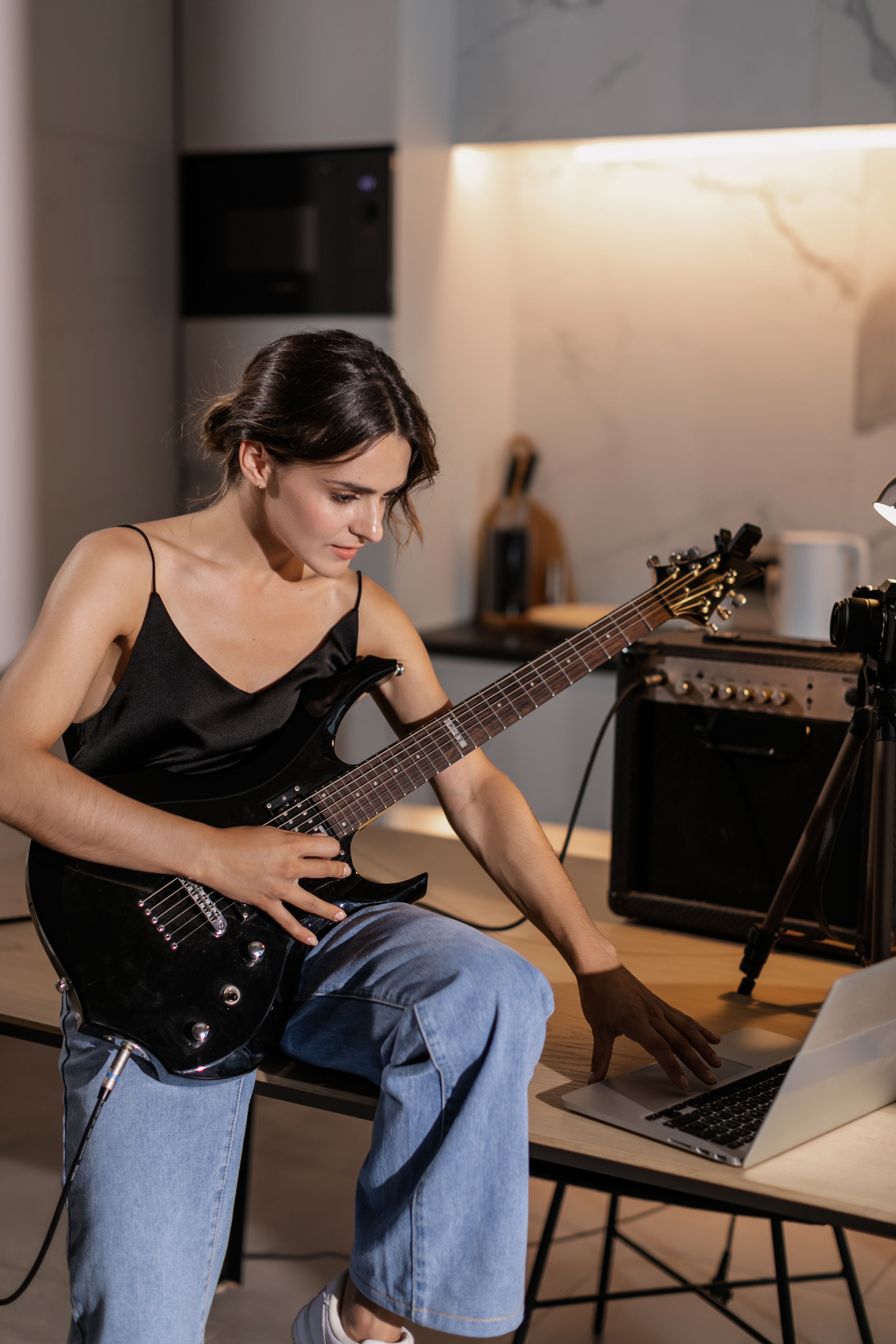 Adult woman playing electric guitar and using laptop in modern home interior.