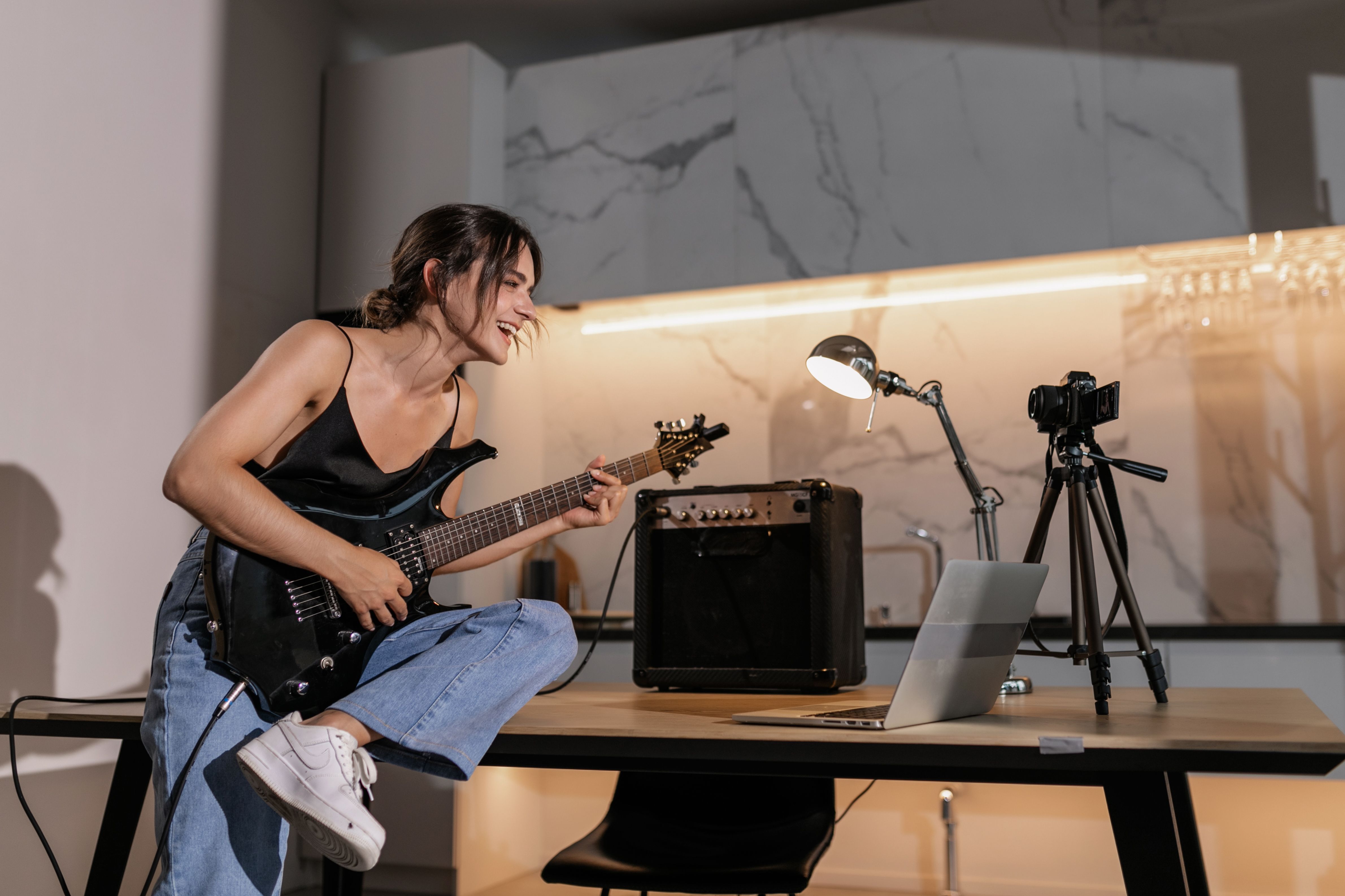 A woman in casual wear playing electric guitar at home, engaging in online learning with a laptop.
